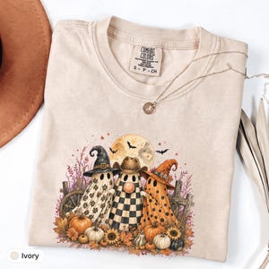 Pode incluir: Uma t-shirt cor de marfim com um gráfico de três fantasmas a usar chapéus, incluindo um chapéu de bruxa preto, um chapéu de cowboy castanho e um chapéu de bruxa laranja. Os fantasmas estão rodeados por abóboras, girassóis e lua cheia.