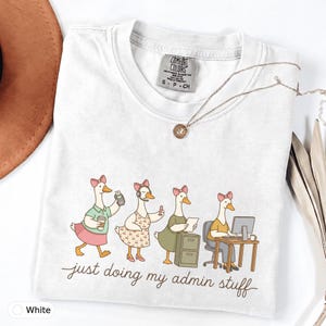 May include: A white t-shirt featuring a graphic of four geese wearing pink bows and performing office tasks, with the text "just doing my admin stuff" printed below. The shirt has a Comfort Colors brand label on the inside of the collar.