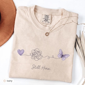 May include: An ivory short-sleeve t-shirt with a graphic print featuring a purple heart, a black line scribble, and a purple butterfly connected by a thin black line. The text "Still Here." is printed below the graphic in a black script font.