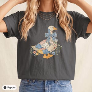 Può includere: Una t-shirt a maniche corte color grigio antracite con una grafica centrale raffigurante un'anatra composta da vari motivi patchwork floreali e a pois. L'anatra indossa un grande fiocco blu intorno al collo ed è circondata da piccoli fiori e foglie.