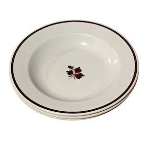 May include: A set of three white ceramic soup bowls with a brown rim and a brown leaf design in the center.