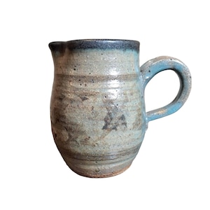 May include: A small, hand-thrown ceramic pitcher with a blue glaze and a brown rim. The pitcher has a rounded body and a small handle.