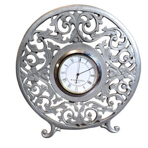 Könnte beinhalten: Eine silberfarbene, verzierte Uhr mit weißem Zifferblatt und schwarzen römischen Ziffern. Die Uhr steht auf einem dekorativen Ständer mit drei Beinen. Das Zifferblatt der Uhr zeigt "Kirk Stief" und "Quartz".