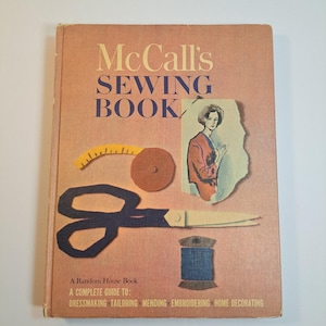 May include: A vintage McCall's Sewing Book with a pink cover featuring a pair of scissors, a spool of thread, a measuring tape, and a woman in a red dress. The book title is "McCall's Sewing Book" and the subtitle is "A Random House Book. A Complete Guide To: Dressmaking, Tailoring, Mending, Embroidering, Home Decorating."