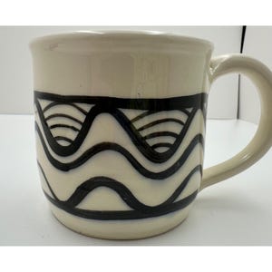 Può includere: Una tazza in ceramica color crema con un motivo dipinto a mano in nero, composto da linee ondulate orizzontali ripetute e archi semicircolari. La tazza presenta una finitura liscia e lucida, un corpo cilindrico largo e un manico curvo.