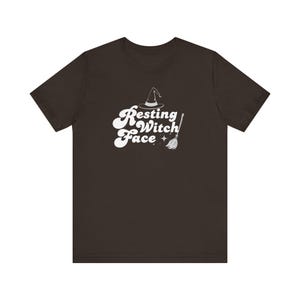 May include: A dark brown short-sleeve t-shirt featuring a white graphic print on the chest. The design includes a witch hat, a broomstick, and the text "Resting Witch Face" written in a bold, wavy, retro-style font with small star accents.