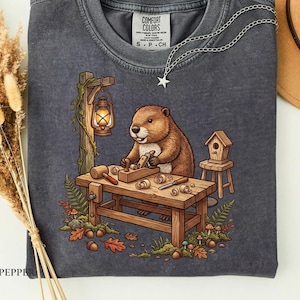 Könnte beinhalten: Ein anthrazitfarbenes T-Shirt mit Rundhalsausschnitt und einer Grafik eines braunen Bibers, der an einer Werkbank aus Holz mit Werkzeugen, einer Laterne und einem Vogelhaus arbeitet. Das Shirt hat ein COMFORT COLORS Etikett und eine silberne Sternkette.