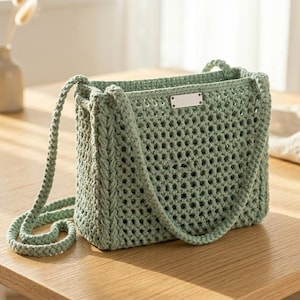 May include: A sage green crochet handbag featuring an open-work mesh pattern, a thick braided shoulder strap, and a rectangular silver-toned metal plate centered on the upper front edge. The bag has a structured rectangular base and sides.