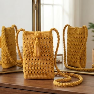 Puede incluir: Un bolso bandolera de ganchillo color amarillo mostaza con cuerpo rectangular de punto texturizado, correa larga trenzada y una borla decorativa central unida a un detalle de nudo en la abertura superior de la pequeña bolsa vertical.
