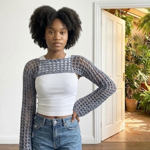 May include: A grey long-sleeved crochet bolero featuring an open-knit grid pattern. The garment is worn over a plain white tank top and blue denim jeans. The bolero has a wide neckline and long sleeves that extend past the wrists, crafted from a textured yarn.