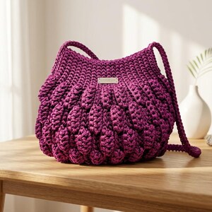 May include: A deep magenta crochet handbag featuring a textured, bobble-stitch pattern throughout the body. The bag includes a matching crocheted shoulder strap and a small, rectangular metallic brand tag centered on the upper front section.