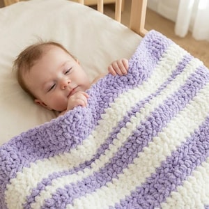 Puede incluir: Una manta de bebé de punto grueso con rayas horizontales alternas de hilo color lavanda suave y crema. La manta tiene un patrón de puntada de bucle grueso y texturizado sobre una cuna de madera con un colchón de color blanco roto.