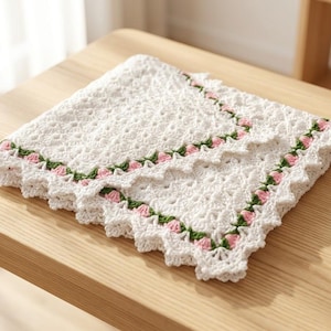 May include: A square white crochet blanket featuring an intricate openwork pattern and a decorative border. The border is embellished with small, hand-stitched pink rosebuds and green leaves, creating a delicate floral accent along the edges.