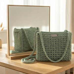 May include: A sage green crochet handbag featuring an open-knit mesh pattern, two sturdy shoulder straps, and a small rectangular metal tag centred on the upper front edge. The bag is constructed from thick yarn with a visible, textured weave.
