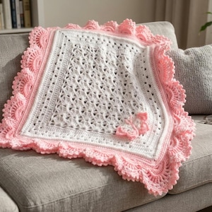 May include: A square crochet blanket featuring a white center with an intricate openwork stitch pattern and a wide, ruffled border in light pink. A small, three-dimensional crocheted butterfly in light pink is attached to the lower right corner.