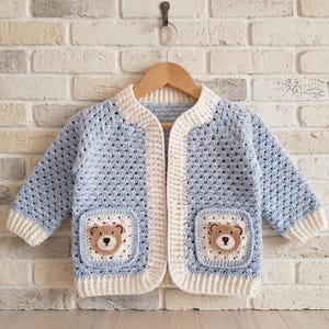 May include: A light blue and cream crochet cardigan featuring long sleeves and two square front pockets. Each pocket displays a small, brown and cream crocheted bear face. The garment has a cream-colored ribbed trim along the neckline, front opening, and sleeve cuffs, and is displayed on a wooden hanger.