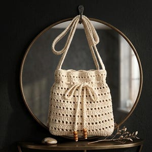 May include: A cream-coloured crochet drawstring bag featuring an open-weave pattern, a long shoulder strap, and a drawstring closure finished with two wooden beads at the ends of the cord. The bag is constructed from yarn in a neutral off-white shade.