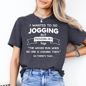 May include: A dark charcoal gray short-sleeve t-shirt featuring white text that reads: I WANTED TO GO JOGGING BUT PROVERBS 28:1 Says THE WICKED RUN WHEN NO ONE IS CHASING THEM. SO THERE'S THAT... The shirt has a crew neckline and a standard fit.