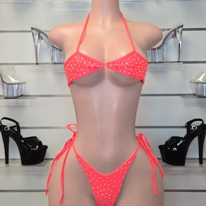 Może przedstawiać: Neonowo-pomarańczowy dwuczęściowy zestaw bikini składający się z trójkątnego topu typu halter oraz dopasowanego dołu wiązanego po bokach. Materiał jest ozdobiony drobnymi, srebrzystymi cyrkoniami rozmieszczonymi na całej powierzchni obu części stroju.