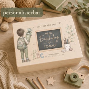 Könnte beinhalten: Eine rechteckige Erinnerungsbox aus hellem Holz mit einer aufgedruckten Illustration eines Kindes mit Rucksack und Schultüte. In der Mitte befindet sich eine Tafelgrafik mit dem Text "MEINE Einschulung TOMMY" sowie "HEUTE IST MEIN TAG!"
