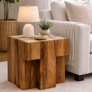 May include: A square wooden side table with a natural grain finish, featuring a thick block-like base. On top sits a white ceramic table lamp with a beige fabric shade, a small potted green plant, and a shallow circular bowl containing small items.