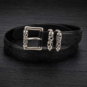 Pode incluir: Um cinto de couro preto com um padrão de cruzes em relevo ao longo da tira. O cinto inclui uma fivela retangular de metal prateado e dois passadores de metal a condizer, todos decorados com intrincados trabalhos de volutas.