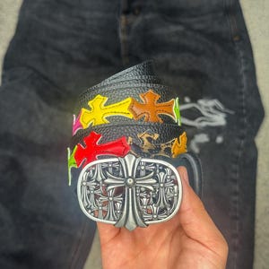 May include: A black textured leather belt featuring a large oval silver-toned buckle with an intricate cross design. The belt strap is decorated with multiple colourful cross-shaped patches in yellow, orange, red, and green, arranged in a row.