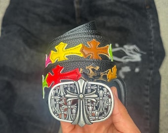 Puede incluir: Un cinturón de cuero negro texturizado con una gran hebilla ovalada de tono plateado con un intrincado diseño de cruz. La correa del cinturón está decorada con varios parches coloridos en forma de cruz en amarillo, naranja, rojo y verde.