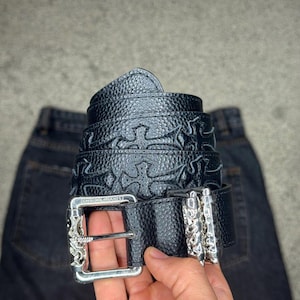 Può includere: Una cintura in pelle nera testurizzata con motivi a croce in rilievo lungo il cinturino. La cintura presenta una fibbia rettangolare in metallo color argento con incisioni dettagliate e la scritta CHROME HEARTS sul telaio superiore.
