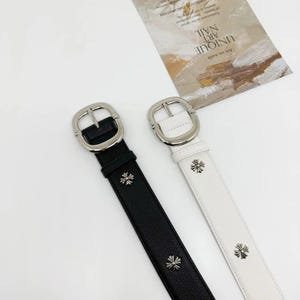 Op de afbeelding: Twee riemen, één zwart en één wit, elk voorzien van een zilverkleurige gesp en gelijkmatig verdeelde zilverkleurige kruisvormige metalen studs op de band. De riemen hebben een gestructureerd oppervlak en liggen naast elkaar op een ondergrond.