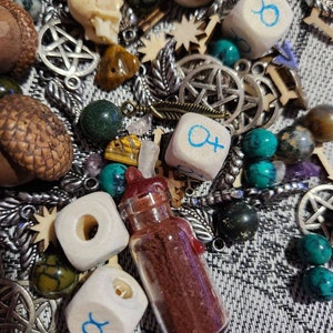 Op de afbeelding: Een verzameling diverse mystieke items, waaronder dobbelstenen met astrologische symbolen, kleine glazen flesjes met rode inhoud, pentagram bedels, houten vormen, eikels en diverse edelstenen. De items zijn gerangschikt op een patroonstof.