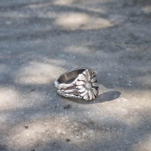 Op de afbeelding: Een zilverkleurige metalen ring met een gedetailleerd, verhoogd zonnebloemontwerp op de voorzijde. De ringband heeft een getextureerd, organisch uiterlijk met gebeeldhouwde blad- en stengelmotieven die om de vinger heen lopen, afgewerkt in gepolijst zilver.