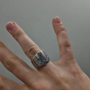 Op de afbeelding: Een brede zilverkleurige metalen ring met een ingewikkeld reliëf bloemmotief over het oppervlak. De band heeft een opvallend, licht onregelmatig randontwerp dat om de vinger wikkelt, met een gepolijste afwerking en donkere verdiepte details.