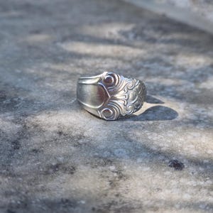 May include: A silver-toned metal ring crafted from a repurposed spoon handle. The band features a smooth, plain shank that transitions into a wide, decorative face adorned with intricate, embossed swirling patterns and floral-inspired motifs.