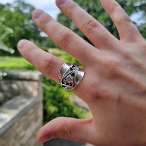 Op de afbeelding: Een zilverkleurige ring gemaakt van een hergebruikte lepel, voorzien van een ingewikkeld reliëf bloemmotief met donkere geoxideerde details in de verdiepte delen van het metaalontwerp, in een brede bandstijl om de vinger.