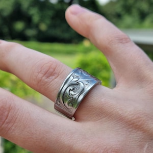 Op de afbeelding: Een brede zilverkleurige metalen ring met een reliëf van bloemrijke krulpatronen met ingewikkelde wervelende lijnen en kleine cirkelvormige accenten. De band heeft een gepolijste afwerking en een plat profiel, dat om de vinger wikkelt met een zichtbare naad.
