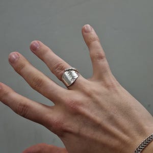 Op de afbeelding: Een zilverkleurige ring gemaakt van een hergebruikte lepel, met een reliëf bloemmotief op de brede band. De ring wikkelt zich om de vinger met een gedraaid handvatontwerp aan de bovenkant en heeft een metallic afwerking met details.