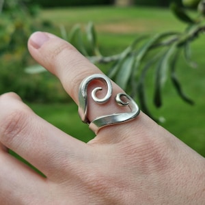 Op de afbeelding: Een zilverkleurige ring gemaakt van een hergebruikt metalen bestekonderdeel, met een opvallend spiraalontwerp dat om de vinger krult. De ringband is gevormd uit het handvat van het bestek en heeft een gladde, gepolijste afwerking.
