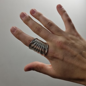 Op de afbeelding: Een brede zilverkleurige ring met een opvallende verticale geribbelde of gegroefde textuur die om de vinger wikkelt. Het metalen oppervlak heeft een gepolijste afwerking met subtiele donkere accenten in de groeven voor een ruimtelijk effect.
