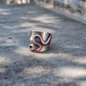 Op de afbeelding: Een brede ring met een basis van donker geoxideerd zilverkleurig metaal, voorzien van een contrasterende, verhoogde, gepolijste koperkleurige draad in een doorlopend, kronkelend, slangachtig of S-vormig patroon rond de buitenkant.
