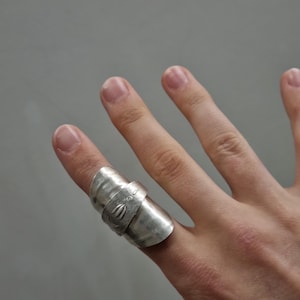 Könnte beinhalten: Ein breiter silberfarbener Ring mit einem Wickeldesign, das an einen gebogenen Löffelstiel erinnert. Die Oberfläche hat ein poliertes metallisches Finish mit einem kleinen eingravierten Blumenmotiv auf dem überlappenden Bandabschnitt.