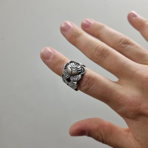 Op de afbeelding: Een zilverkleurige metalen ring met een gedetailleerd, driedimensionaal roosontwerp. Het bloemmotief bedekt het bovenste gedeelte van de ring en toont gebeeldhouwde bloemblaadjes en bladeren met donkere uitsparingen om het metaalwerk te accentueren.