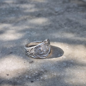 Op de afbeelding: Een zilverkleurige metalen ring gemaakt van een hergebruikte lepel, met een sierlijk reliëfpatroon van krullen op de voorzijde en een textuurafwerking. De binnenkant van de ring toont een duidelijk keurmerk 1847 Rogers Bros.