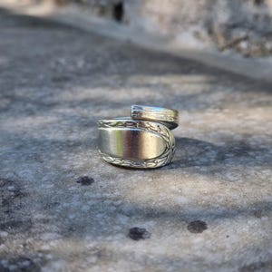 May include: A silver-toned metal ring crafted from a repurposed spoon, featuring a bypass design where the handle and bowl ends overlap. The band displays an embossed floral or vine pattern along the edges, with a smooth, polished centre section.