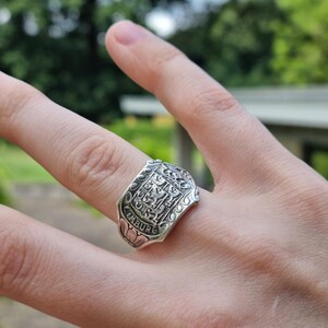 Op de afbeelding: Een zilverkleurige ring met een reliëf van een heraldisch schildontwerp met een leeuwembleem en de tekst LIMBURG onderaan. De ring heeft een brede, platte voorzijde met decoratieve kruldetails langs de randen en een gepolijste afwerking.