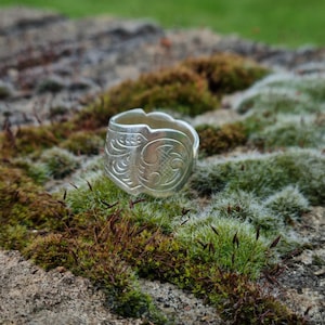 Könnte beinhalten: Ein Silberring, gefertigt aus einem umfunktionierten Löffel, mit einem komplizierten geprägten Blumen- und geometrischen Muster auf dem äußeren Band. Das Metall hat eine polierte Oberfläche mit sichtbaren dekorativen Gravuren rundherum.
