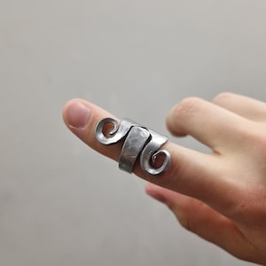 Op de afbeelding: Een zilverkleurige metalen ring met een gehamerde textuur en een ontwerp met twee symmetrische, naar buiten gerichte spiralen aan weerszijden van een centrale band. Het metaal heeft een licht matte, industriële afwerking met zichtbare sporen.