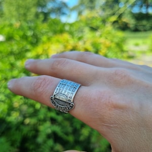 May include: A silver-toned metal ring featuring a detailed embossed design of a building facade with multiple windows and a roofline. The base of the ring displays the text BIJENKORF in raised lettering, curved along the bottom edge of the band.