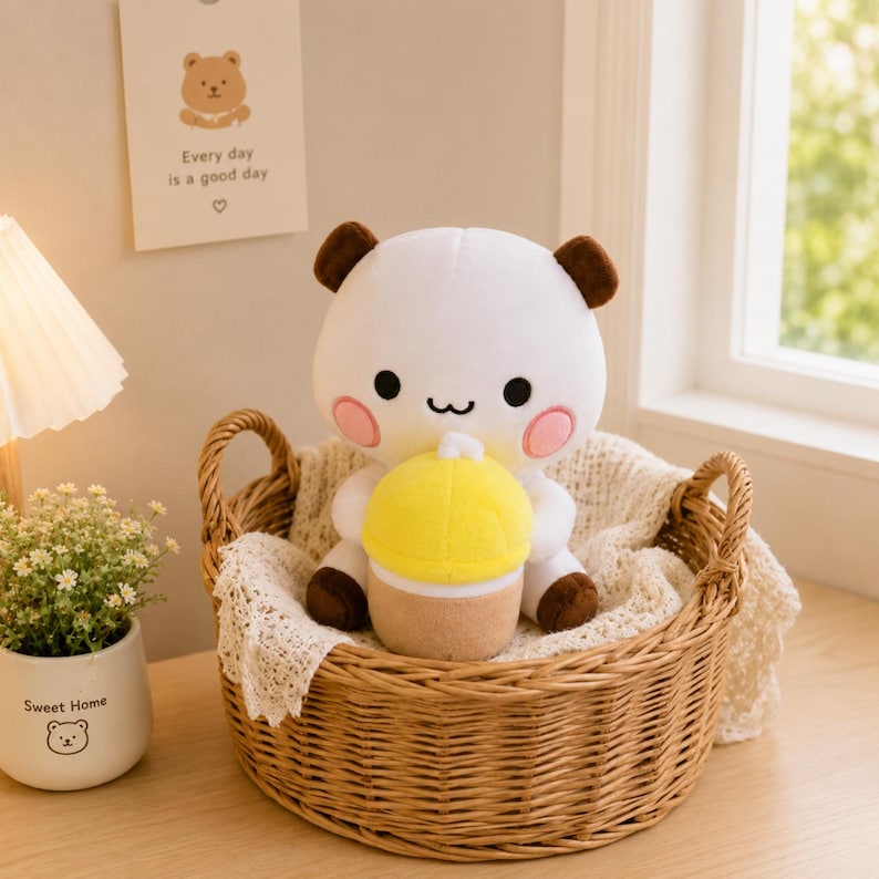 May include: A white plush bear toy with brown ears and pink cheeks holding a yellow and tan cupcake. The bear has black eyes and a small black mouth. It is sitting inside a round woven wicker basket lined with a white lace-trimmed fabric.