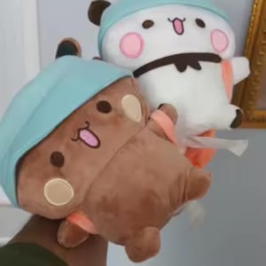 May include: Two plush toys featuring rounded heads and bodies. One toy is brown with a light blue hat and peach-colored cheeks, while the other is white with a light blue hat, pink cheeks, and a black bow tie. Both have embroidered faces.